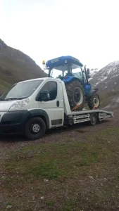 Uygun fiyatlı ve acil yol yardımı hizmetlerini temsil eden ve arazide taşınan traktör görseli.